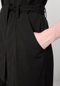 Vestido de tela negra con una cintura definida, bolsillos laterales y un cinturón anudado. Textura suave con un diseño aerodinámico y detalles de costura mínimos.