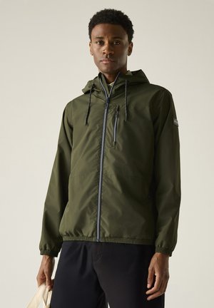Uomo che indossa una giacca con cappuccio verde oliva con cerniera e pantaloni neri, tenendo una borsa bianca su uno sfondo semplice.