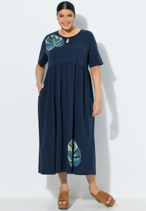 Lächelnde Frau im marineblauen Midikleid mit kurzen Ärmeln, Ausschnitt mit Schlitz, zwei grünen Blattmustern und beige Plateau-Sandalen.