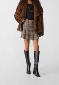Bruine nepbontjas, zwarte getailleerde top, beige minirok met rode lijnen, en hoge zwarte leren haklaarzen.