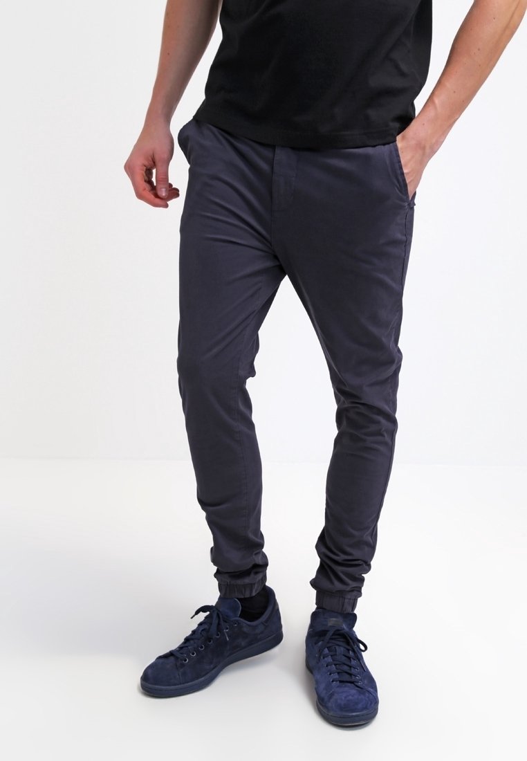 Mörkgrå tapered byxor har en elastisk midja, sidofickor och stretchigt material för komfort. De är matchade med marinblå sneakers.