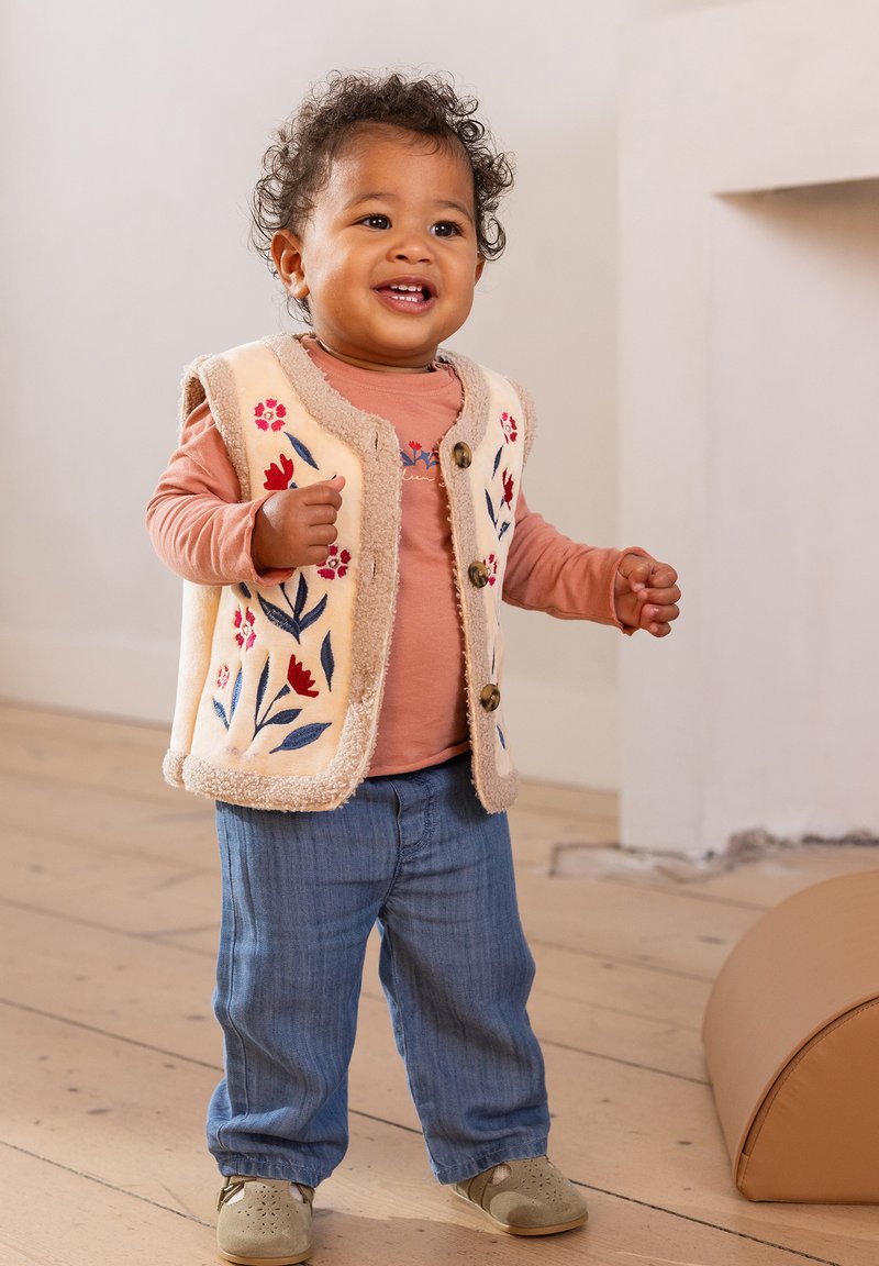 Korte mouwen roze shirt onder een crème, fluffy vest met bloemenborduursel, gecombineerd met lichtblauwe spijkerbroek en beige schoenen.