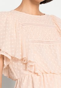 Blouse pêche clair en tissu à pois suisses texturé, avec une bordure en dentelle sur la poitrine et des manches flottantes superposées, portée par une personne.