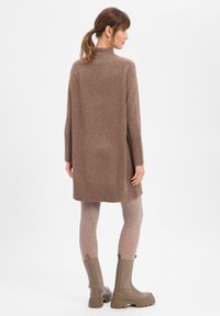 Kobieta widziana od tyłu, ubrana w długi brązowy sweter, beżowe legginsy i masywne brązowe skórzane buty, stojąca na białym tle.