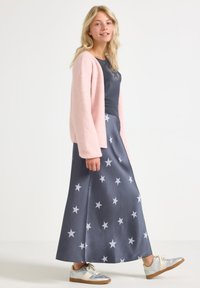 Femme aux cheveux blonds portant un cardigan rose doux, une jupe bleu marine à motifs d'étoiles, un haut foncé avec un graphisme d'étoile et des baskets argentées marchant de côté.