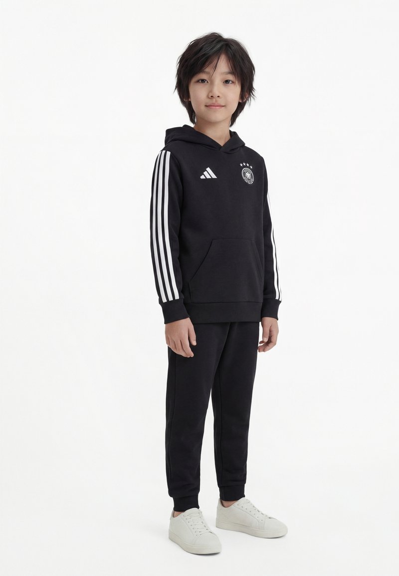Niño de pie vistiendo ropa deportiva Adidas negra con rayas blancas y zapatillas blancas contra un fondo blanco liso.