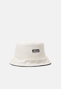 Bucket hat made of cream-colored fabric with a rounded crown, wide floppy brim, and a black rectangular logo patch on the front.