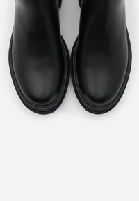 Pár černých kožených bot s kulatými špičkami a dekorativním prošitím na podešvích, zobrazených z pohledu shora na bílém pozadí.