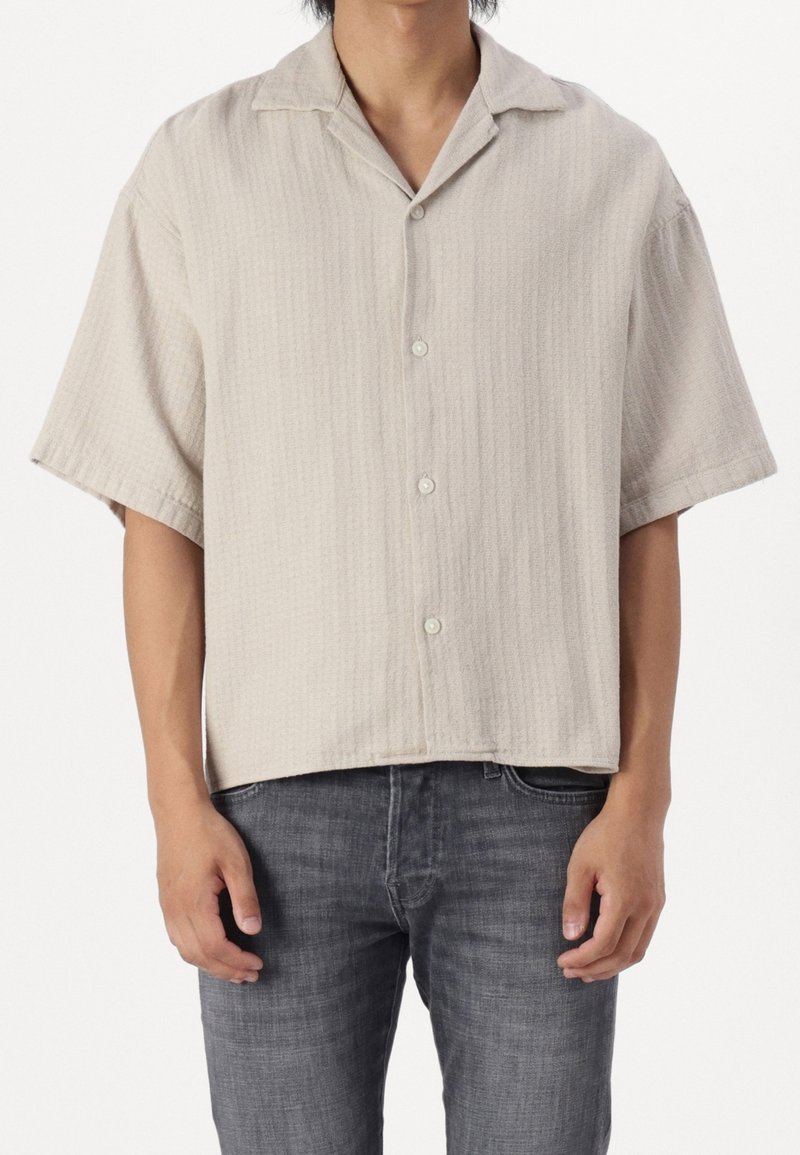 Uomo che indossa una camicia beige a maniche corte e con colletto, larga e con bottoni, insieme a jeans grigio scuro, su uno sfondo bianco.