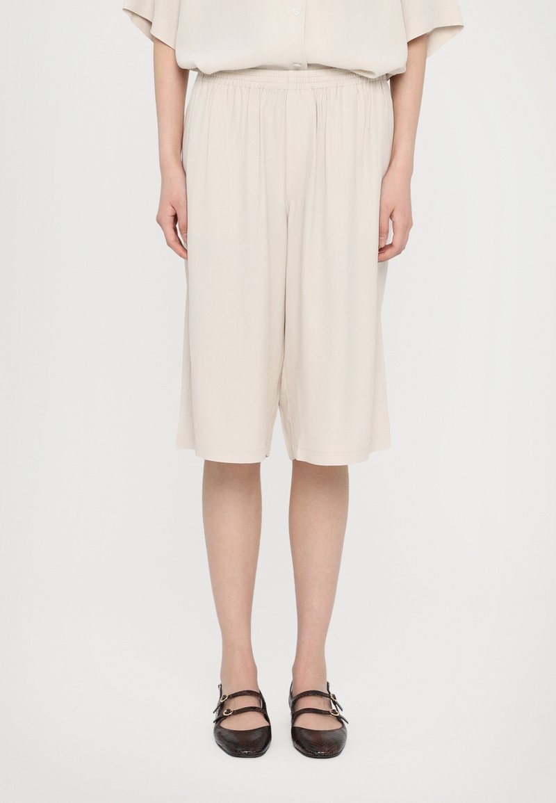 Personne portant un short ample beige clair jusqu'aux genoux, assorti d'un haut similaire et de chaussures Mary Jane noires texturées avec deux brides à boucles.