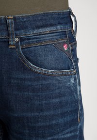 Jeans de mezclilla azul oscuro con un corte clásico, que presentan costuras naranjas y un pequeño diseño de rosa bordada en el bolsillo delantero.