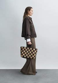 Sac cabas tissé présentant un motif à carreaux marron foncé et beige, avec une anse robuste et une forme rectangulaire spacieuse.