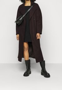 Dlouhý, tmavě purpurový kabát s klopovým límcem, bočními kapsami a hladkým povrchem, kombinovaný s černými šaty a černými kozačkami po kolena.