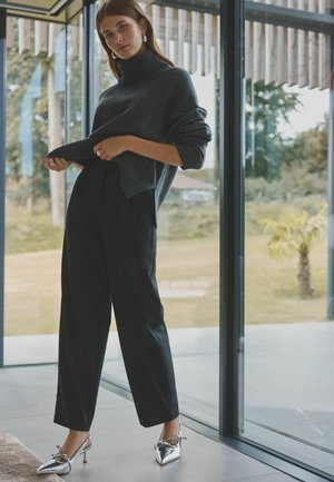 Maglione grigio a costine con collo a imbuto, pantaloni neri a vita alta e décolleté argentati a punta con cinturino posteriore. Design minimalista.