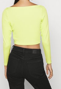 Długi rękaw, krótki top w neonowej żółci, zestawiony z czarnymi jeansami o wysokim stanie. Top ma kwadratowy dekolt i prążkowaną fakturę.