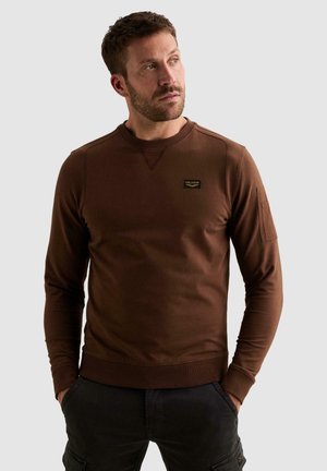 Homme aux cheveux courts et bruns portant un sweat-shirt marron à manches longues et un pantalon foncé, debout les mains dans les poches, regardant vers sa gauche.