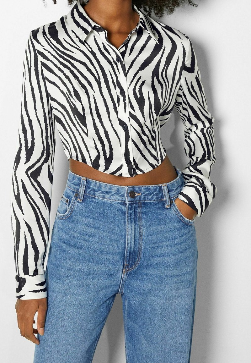 Femme portant une chemise courte à boutons à rayures zébrées noires et blanches, associée à un jean bleu taille haute, main dans la poche.