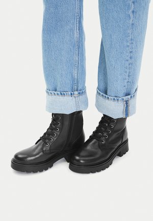 Czarne skórzane buty sznurowane z teksturowanym wykończeniem, grubymi podeszwami i zamkiem błyskawicznym po bokach. W zestawieniu z podwiniętymi jasnoniebieskimi dżinsami.