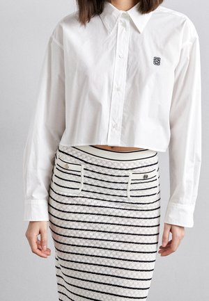 Hvid kort, knaplukket skjorte med lange ærmer og et sort logo på brystet, kombineret med en strikket nederdel med sorte, vandrette striber og lommer.