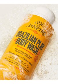 Żółta butelka Sol de Janeiro Brazilian Play Body Wash częściowo pokryta pęcherzykami mydła na białej, mydlanej powierzchni.