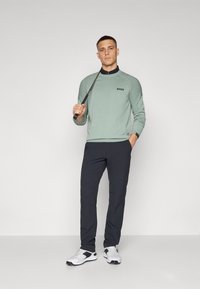 Lys grønn langermet polo med svart logo, kombinert med mørk marinebukser og hvite golfsko med svarte detaljer. Holder en golfsjef.