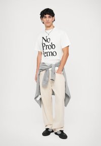 White T-shirt with black print, light gray shirt tied at the waist, beige pants, and black shoes with white stripes. Simple and casual design.