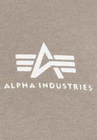 Beige szövet, amelyen egy fehér logó látható, amely egy 'A'-t és vonalakat kombinál, alatta a "ALPHA INDUSTRIES" felirat található. Texturált felület.