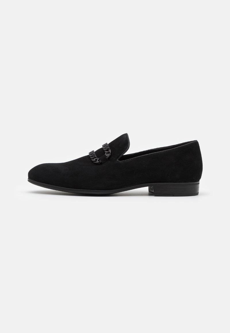 Mocassin en daim noir pour homme avec un détail de chaîne tordue sur le dessus et un petit talon empilé sur un fond uni.