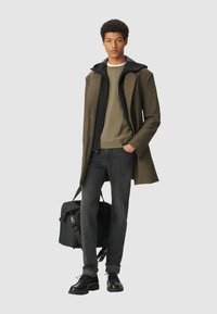 Joven de pie vestido con un abrigo verde y negro en capas, suéter oliva, jeans oscuros, zapatos negros, sosteniendo un bolso de deporte negro.