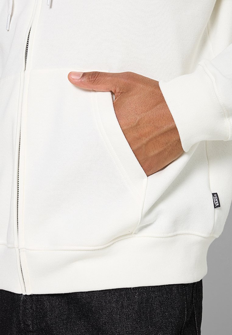 Biała bluza z kapturem na zamek błyskawiczny, z kieszenią kangurzą, ściągaczami przy mankietach i małą czarną metką po boku, prezentowana na neutralnym tle.
