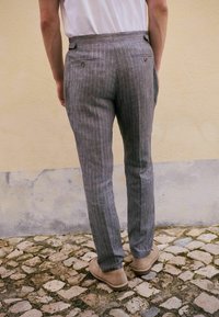 Pantalon gris à rayures fines en tissu léger avec deux poches arrière et des accents de boutons, porté avec des chaussures beiges sur des pavés.
