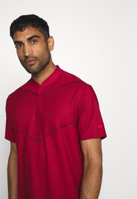 Röd sportpolo med en subtil chevrondesign, texturerad tyg och mandarinkrage. Har en broderad logotyp på ärmen.