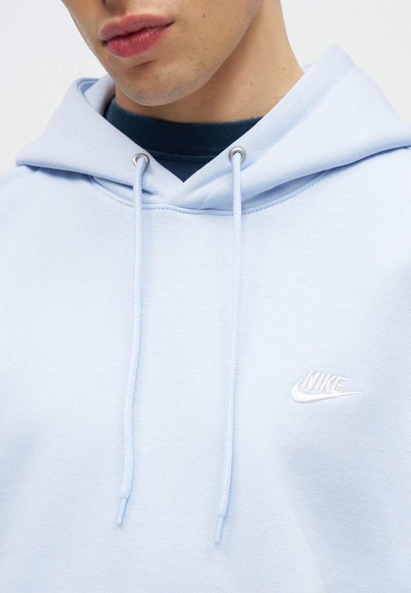 Primer plano de una persona que lleva una sudadera con capucha Nike de color azul claro con cordones y un logo blanco de Nike visible en el pecho.