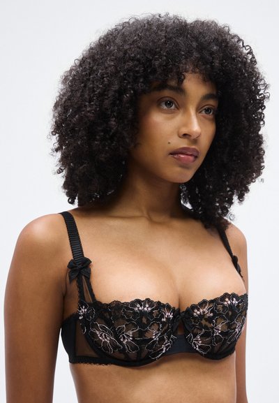 Svart balconette-bh i spets med blommig broderi och genomskinlig mesh. Axelband prydda med små svarta rosetter. Justerbar stängning bak.