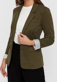 Olivgrön blazer med en knapp, slagkragar och en bröstficka. Veckade ärmar med ljusa randiga detaljer. Bärs över en vit topp.