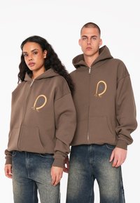 Brauner Zip-Hoodie mit großen vorderen Taschen, ausgestattet mit goldener Stickerei und lässigem Schnitt. Trägt dazu blaue Jeans.