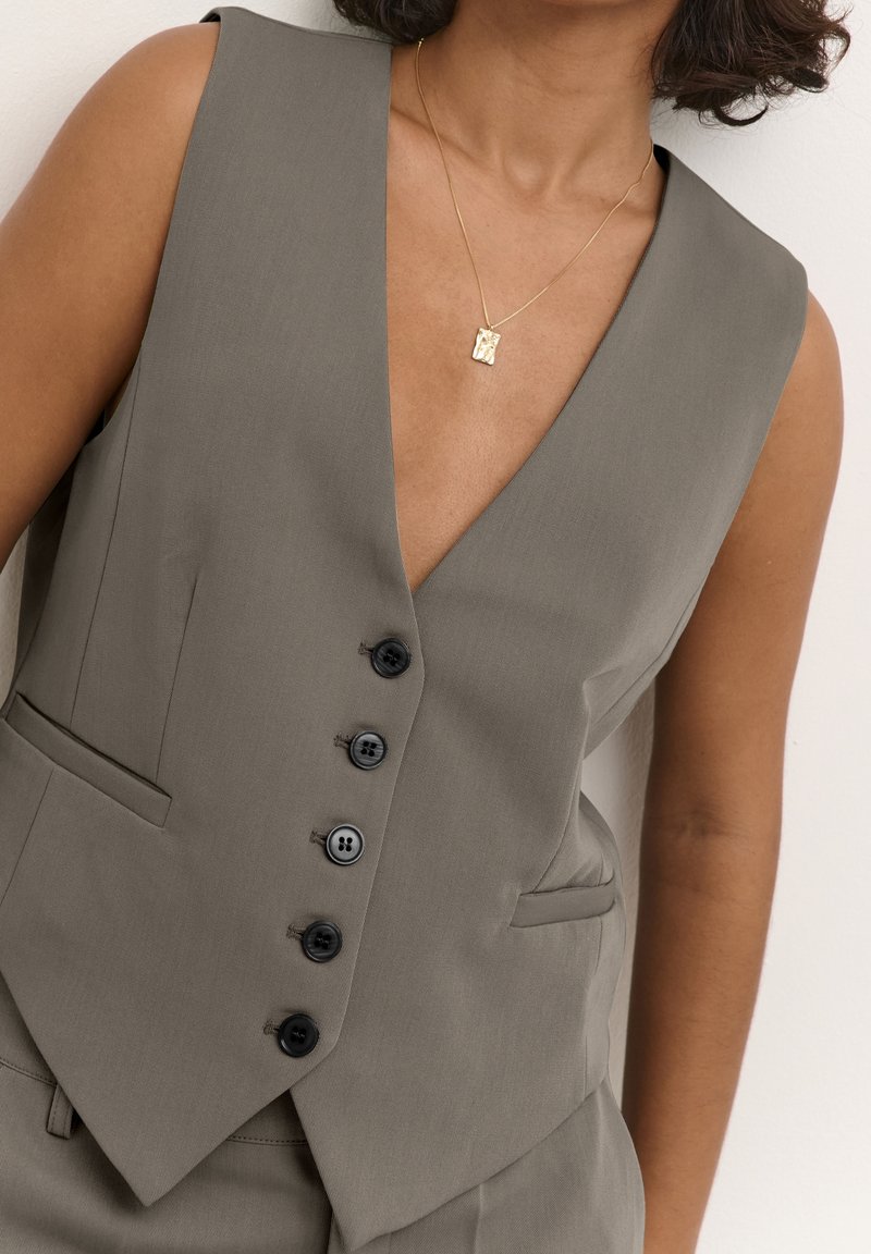 Femme portant un gilet gris sans manches boutonné et un collier pendentif rectangulaire en or, cadrée des épaules à la taille.