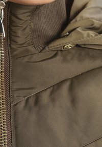 Chaqueta acolchada de color verde oliva con textura acolchada, cuenta con cierre de cremallera y broches, cuello acanalado y forro suave.