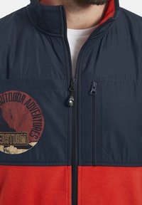 Man in een rood en marineblauwe jas met een ritzak en het logo "Outdoor Adventures" op de borst, gedeeltelijk dichtgeritst over een wit shirt.