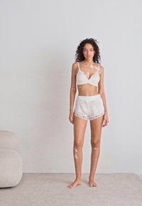 Lichtbeige bralette en bijpassende shorts met geschulpte kantafwerking. Model staat blootsvoets tegen een neutrale muur, eenvoudige achtergrond.