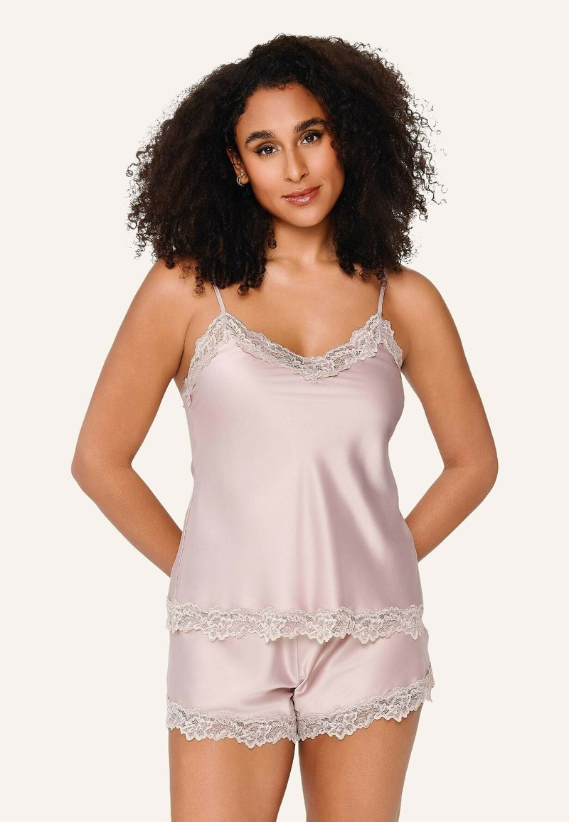 Femme aux cheveux bouclés portant un caraco en satin rose clair et un short assorti avec bordure en dentelle, debout devant un fond uni.