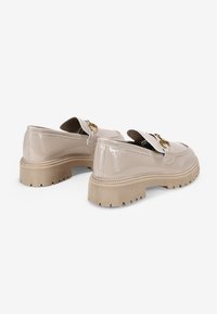 Mocasines de charol beige con punta redonda, diseño deslizante y un prominente detalle en tono dorado en la parte superior. Suela gruesa y texturizada.