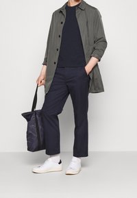 Lätt olivgrön jacka med svarta knappar, marinblå t-shirt, mörk marinblå byxor, vita sneakers och en mörk totes väska. Släta texturer.