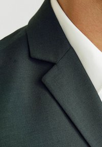 Sötétzöld zakó texturált anyagból, éles hajtókákkal és kontrasztos fehér inggallérral.