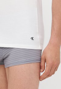 Weißes Baumwoll-T-Shirt mit einem schwarzen Logo am Saum, kombiniert mit grau gestreiften Boxershorts. Nahaufnahme einer Hand, die auf dem Oberschenkel ruht.