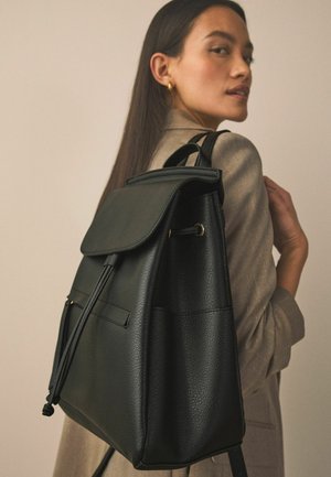 Musta nahkainen reppu, jossa on läppäkansi, nyöritysdetaili ja kullanväriset metalliosat. Pinnan tekstuuri ja kaksi ulkopuolista taskua.