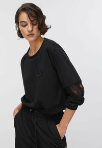 Sweat-shirt noir avec un col rond et des manches longues ornées d'accents en dentelle aux poignets. Logo brodé sur le devant.
