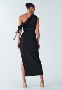 Robe noire à une épaule, avec un tissu plissé, une fente sur le côté et des détails à cordon au niveau des manches et de la taille. Texture lisse, longueur midi.