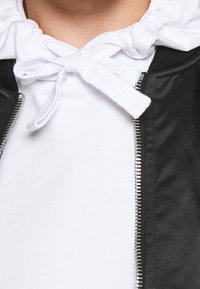 Černá bunda s kovovým zipem, pod kterou je bílá mikina s kapucí a šňůrkami. Detailní záběr, důraz na texturu látky a detaily.