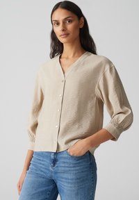 Beige, crinkled fabric shirt with a V-neck, front buttons, and three-quarter sleeves with elastic cuffs. Worn with blue jeans.